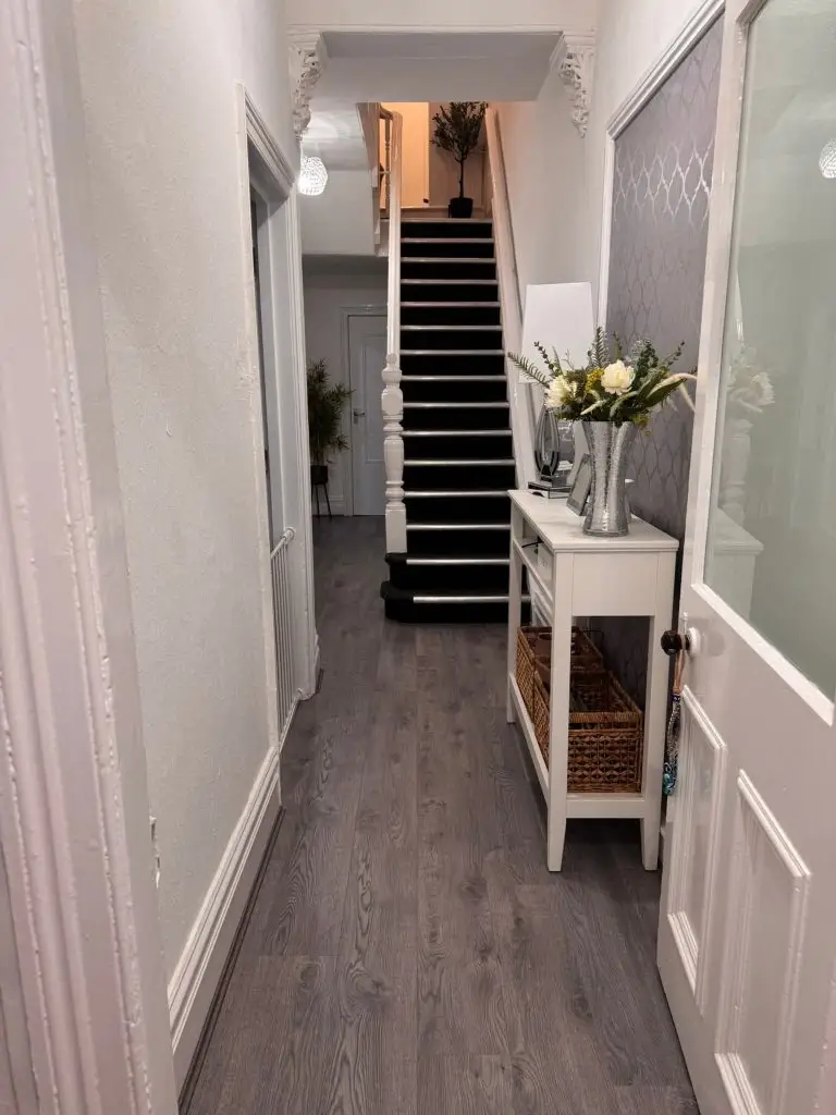 Modern hallway with dark staircase, white console table with flowers, and gray wood-look flooring.