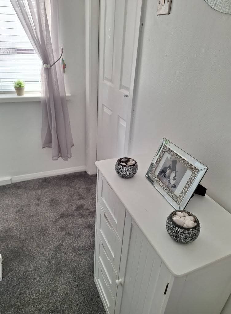 Modern bedroom entryway with white furniture, decorative bowls, framed photo, purple curtains, and gray carpet flooring.