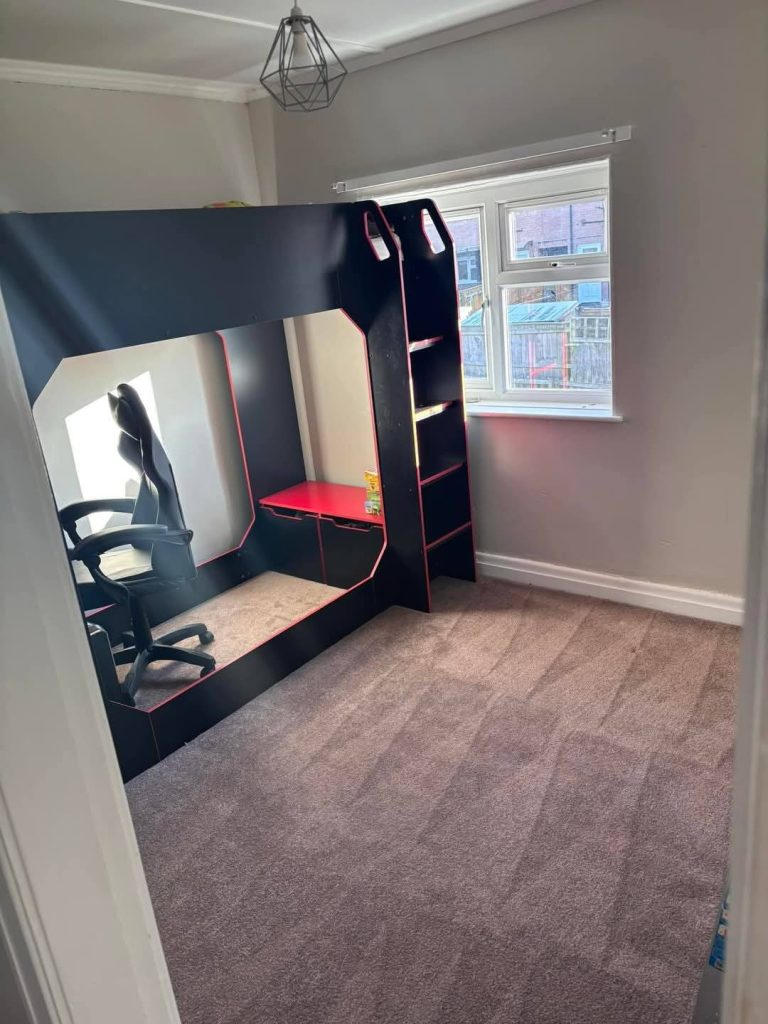 Modern gaming setup with black and red gaming chair, monitor, and shelving unit in bright bedroom with windows.