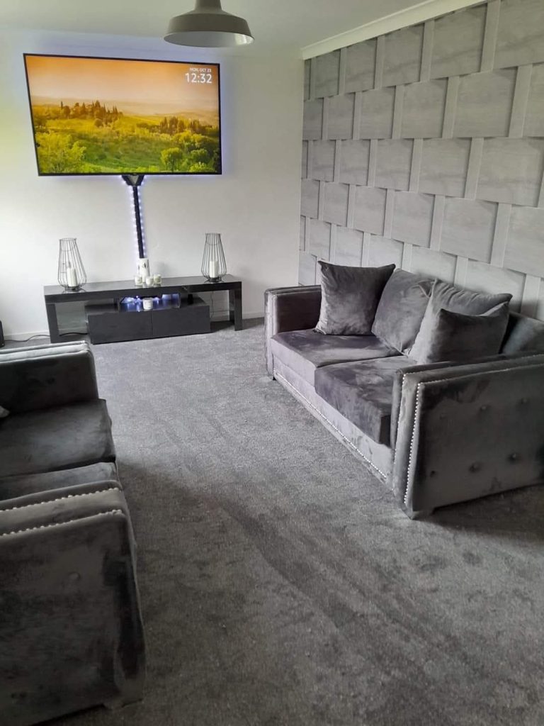 Modern living room with gray upholstered sofa, mounted TV displaying landscape, black media console, and textured accent wall