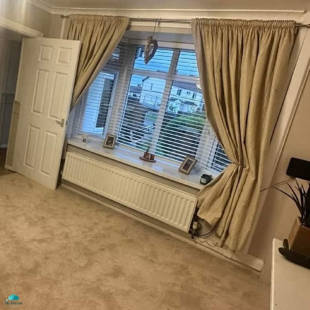 Bright modern living room with large bay window, beige curtains, radiator, and city view through horizontal blinds.