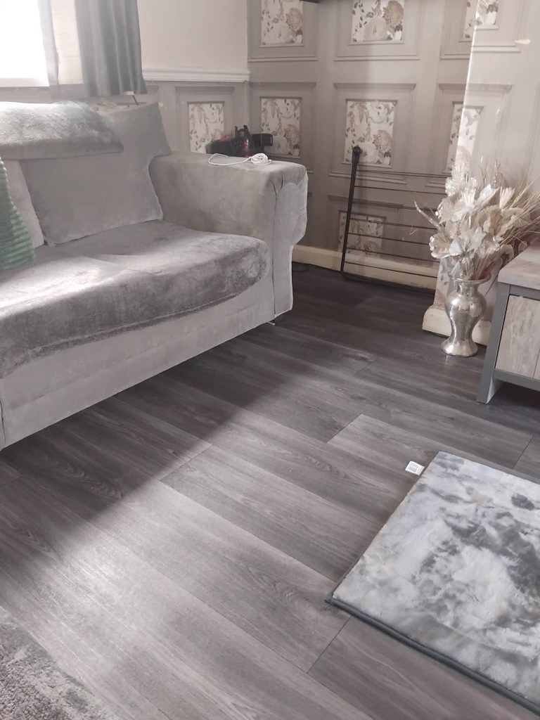 Modern bedroom interior with gray upholstered sofa, dark wood-look flooring, floral wall panels, and white decorative vase with dried flowers.