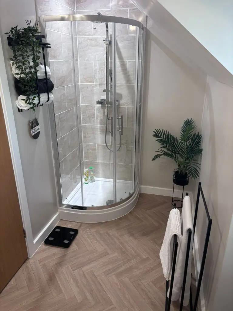 Modern bathroom with curved glass shower enclosure, gray tile walls, herringbone wood flooring, and decorative plants.